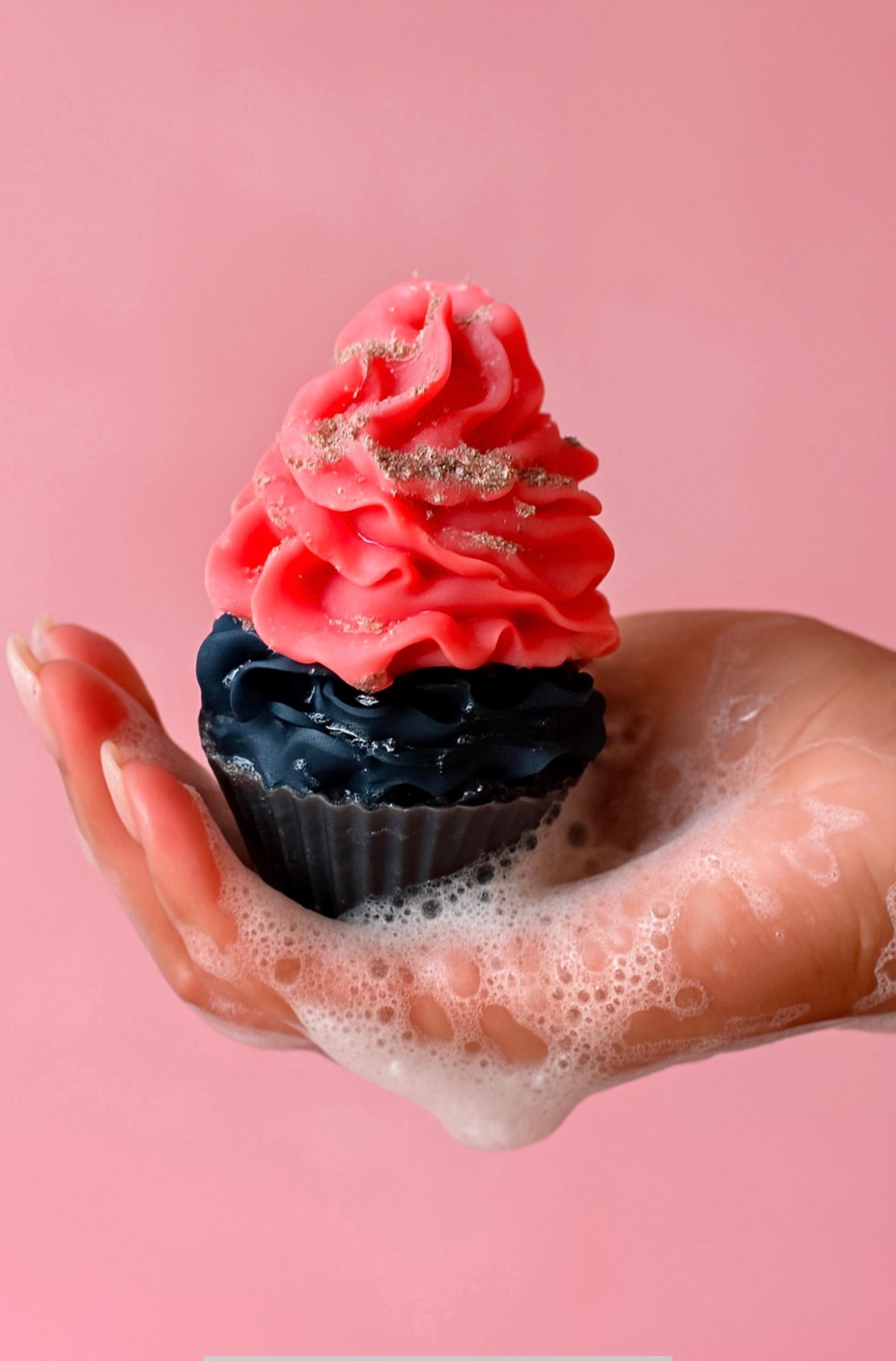 Hand holding a cupcake-shaped soap with a activated charcoal base and pink frosting top, sprinkled with grey sprinkles of activated charcoal and surrounded by light soap bubbles, against a pastel pink background.